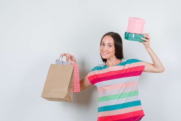 Mujer joven mostrando bolsas de papel y cajas de regalo en camiseta y mirando feliz, vista frontal.