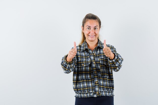 Mujer joven mostrando doble pulgar hacia arriba en camisa, pantalones cortos y mirando alegre. vista frontal.