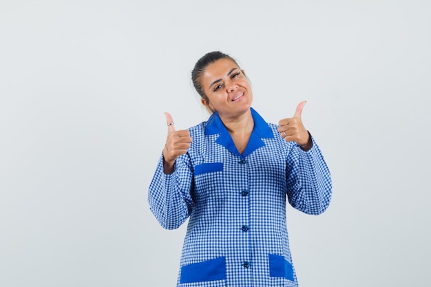 Mujer joven mostrando doble pulgar hacia arriba en camisa de pijama de cuadros azules y luciendo bonita. vista frontal.