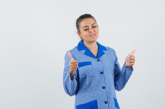 Mujer joven mostrando doble pulgar hacia arriba en camisa de pijama de cuadros vichy azul y mirando serio, vista frontal.