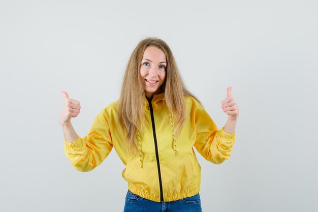 Mujer joven mostrando doble pulgar hacia arriba en chaqueta de bombardero amarilla y jean azul y luciendo optimista, vista frontal.