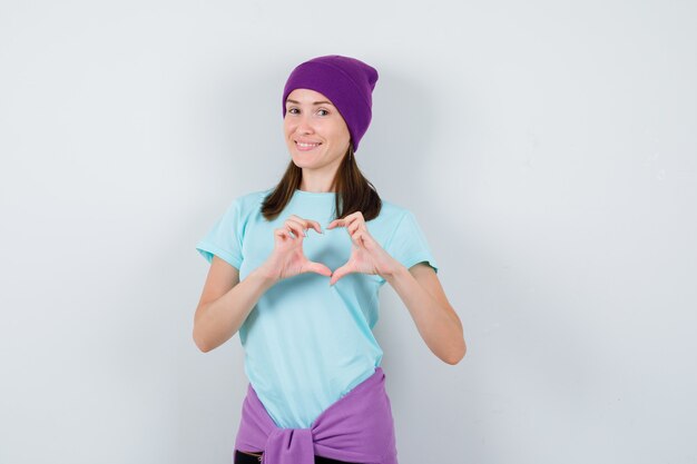 Mujer joven mostrando gesto de corazón en camiseta azul, gorro morado y mirando alegre, vista frontal.