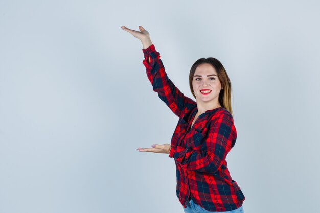 Mujer joven mostrando gesto de escalas en camisa a cuadros y mirando alegre. vista frontal.