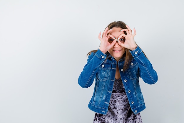 Mujer joven mostrando gesto de gafas en chaqueta de mezclilla, vestido y mirando positivo, vista frontal.