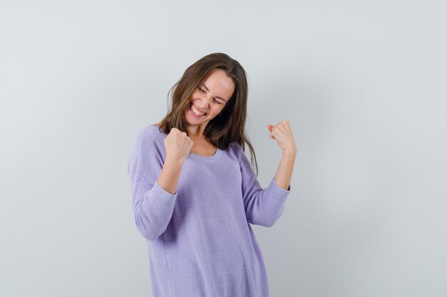 Mujer joven mostrando gesto ganador en blusa lila y mirando alegre