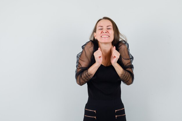 Mujer joven mostrando gesto ganador en blusa negra y mirando alegre