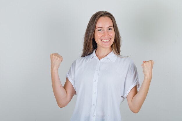Mujer joven mostrando gesto ganador en camisa blanca y mirando feliz