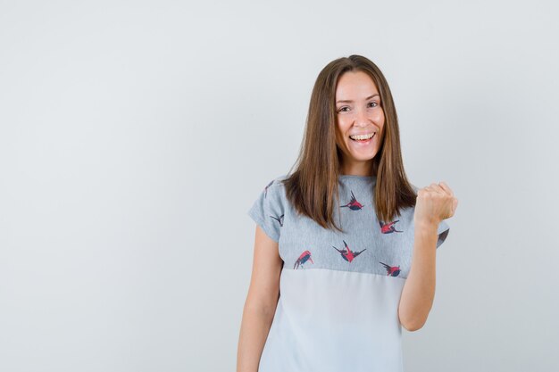 Mujer joven mostrando gesto ganador en camiseta y mirando dichoso, vista frontal.