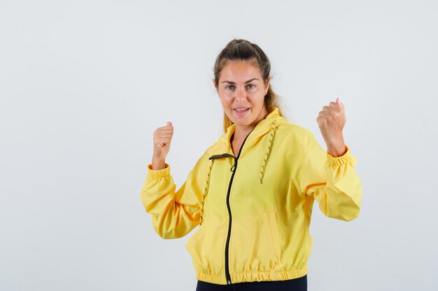 Mujer joven mostrando gesto de ganador en impermeable amarillo y mirando alegre