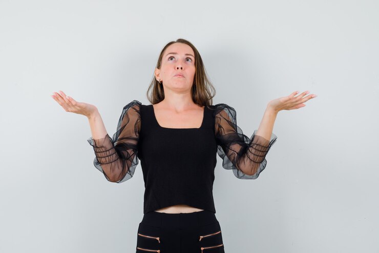 Foto gratuito mujer joven mostrando gesto de impotencia en blusa negra y mirando confundido