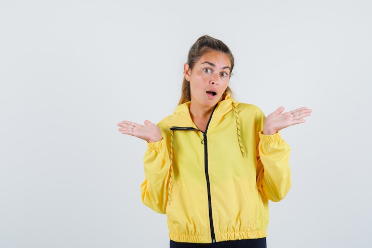 Foto gratuito mujer joven mostrando gesto de impotencia en impermeable amarillo y mirando perplejo