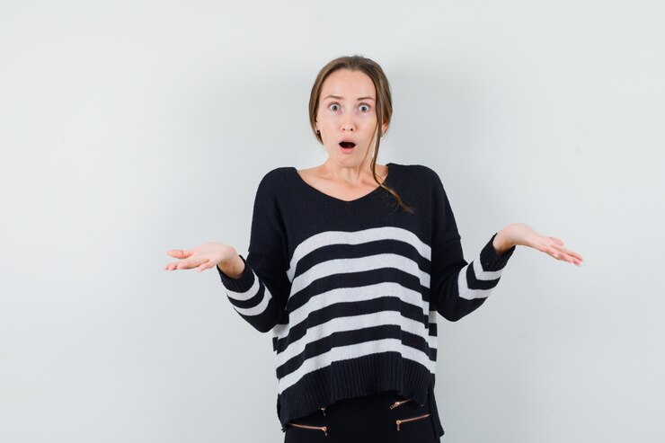 Foto gratuito mujer joven mostrando gesto de impotencia en prendas de punto a rayas y pantalón negro y mirando sorprendido