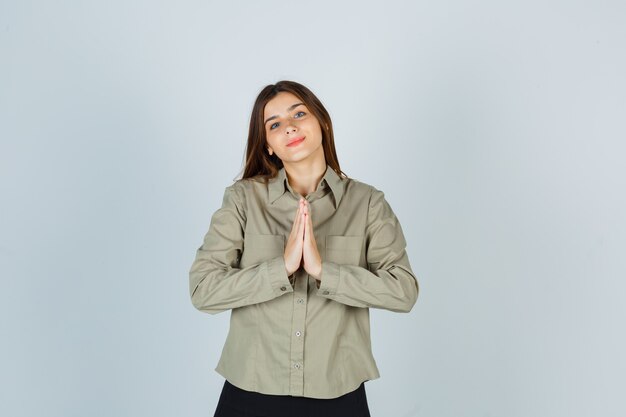 Mujer joven mostrando gesto de namaste en camisa, falda y mirando esperanzado, vista frontal.