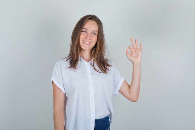 Mujer joven mostrando gesto ok en camiseta blanca, jeans y mirando alegre