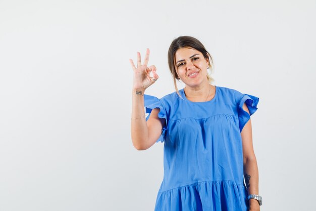 Mujer joven mostrando gesto ok en vestido azul y mirando alegre