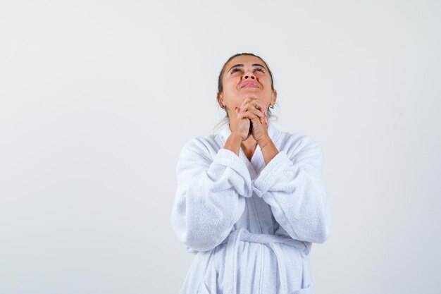 Mujer joven mostrando gesto de oración en bata de baño y mirando esperanzado