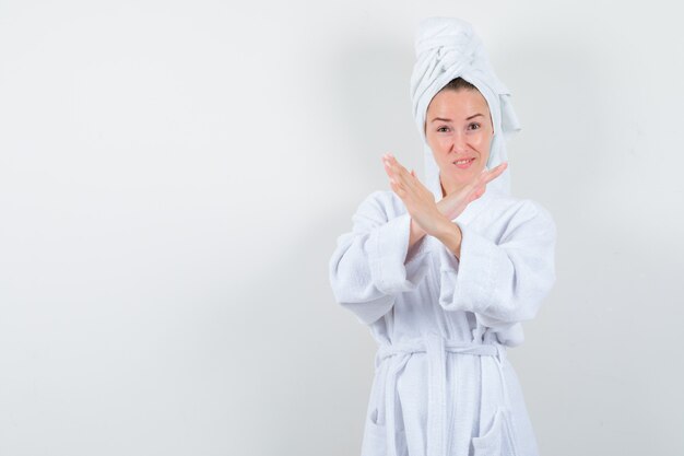 Mujer joven mostrando gesto de parada en bata de baño blanca, toalla y mirando confiado, vista frontal.