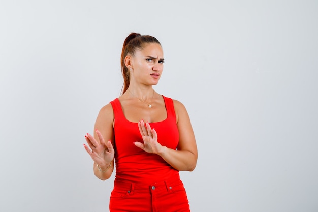 Mujer joven mostrando gesto de parada en camiseta roja, pantalones y mirando disgustado, vista frontal.