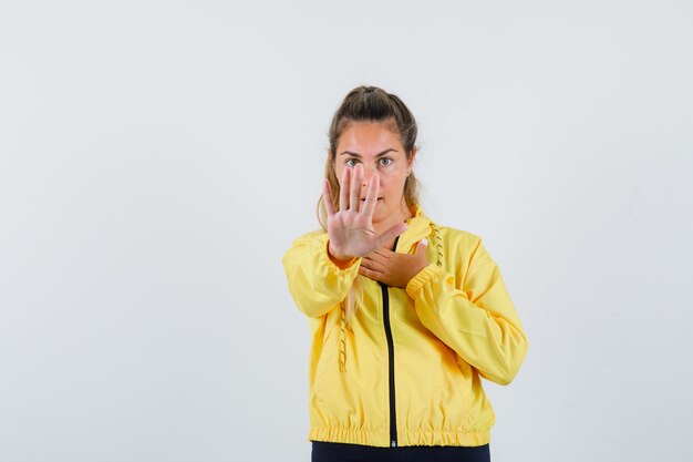 Mujer joven mostrando gesto de parada en impermeable amarillo y mirando reacio