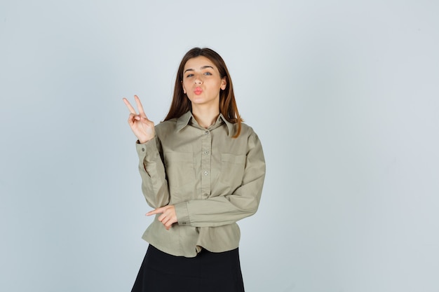 Mujer joven mostrando gesto de paz, haciendo pucheros en camisa, falda y luciendo orgullosa