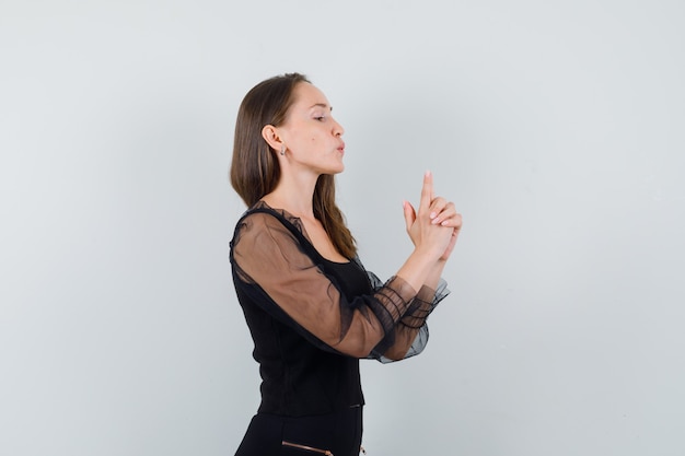Mujer joven mostrando gesto de pistola en blusa negra y pantalón negro y mirando confiado, vista frontal.