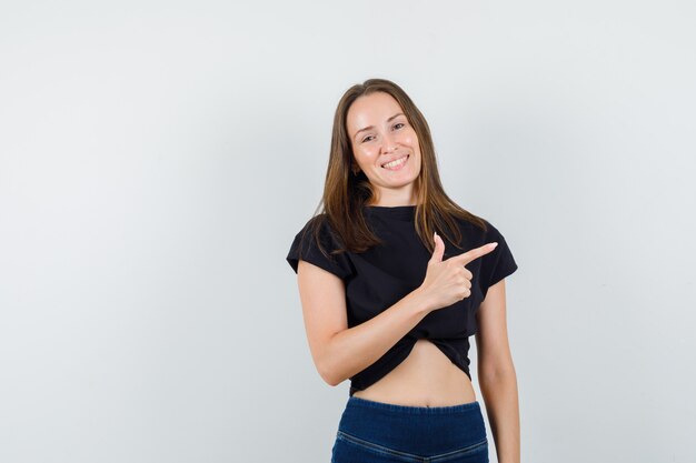 Mujer joven mostrando gesto de pistola en blusa negra, pantalones y mirando feliz