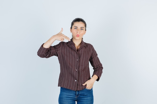 Foto gratuita mujer joven mostrando gesto de pistola, con la mano en el bolsillo en camisa a rayas, jeans y luciendo lindo. vista frontal.