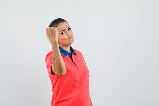 Mujer joven mostrando gesto de poder en camiseta roja y mirando confiado. vista frontal.