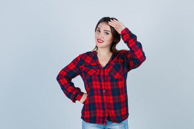 Mujer joven mostrando gesto de saludo en camisa a cuadros y mirando alegre, vista frontal.