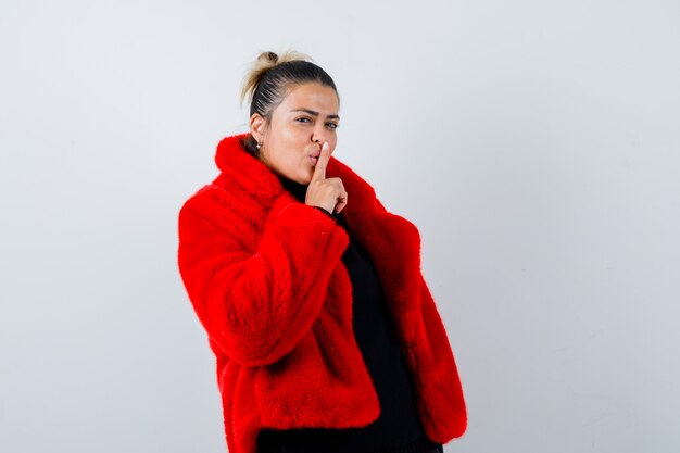 Mujer joven mostrando gesto de silencio en suéter, abrigo de piel rojo y mirando confiado, vista frontal.