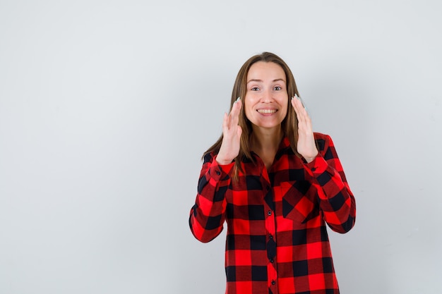 Mujer joven mostrando gesto de tamaño en camisa a cuadros y mirando alegre. vista frontal.