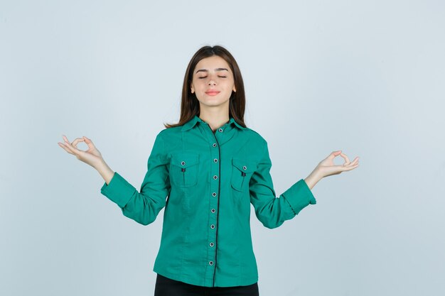 Mujer joven mostrando gesto de yoga con los ojos cerrados en camisa verde y mirando relajado, vista frontal.