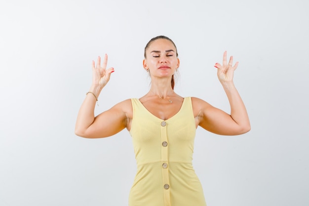 Mujer joven mostrando gesto de yoga con los ojos cerrados en vestido amarillo y mirando relajado. vista frontal.