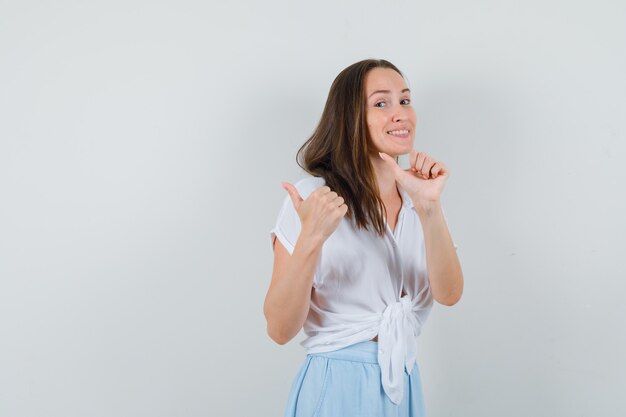 Mujer joven mostrando golpe hacia arriba y apuntando a la izquierda en blusa blanca y falda azul claro y mirando feliz