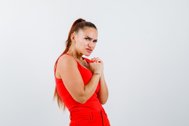 Mujer joven mostrando las manos juntas en gesto de súplica en camiseta roja, pantalones y mirando esperanzado, vista frontal.