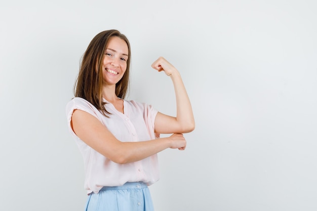 Mujer joven mostrando los músculos del brazo en camiseta, falda y luciendo alegre. vista frontal.