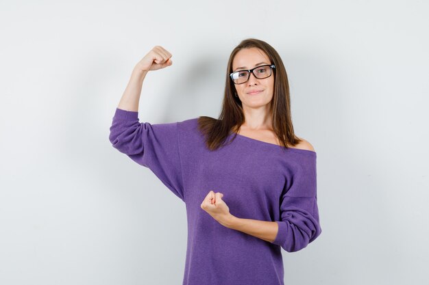 Mujer joven mostrando músculos y puños en camisa violeta y mirando confiada. vista frontal.