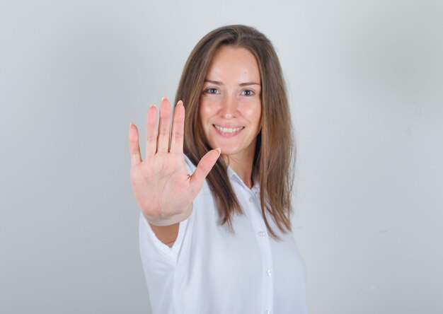 Mujer joven mostrando la palma a la cámara en camiseta blanca y mirando alegre