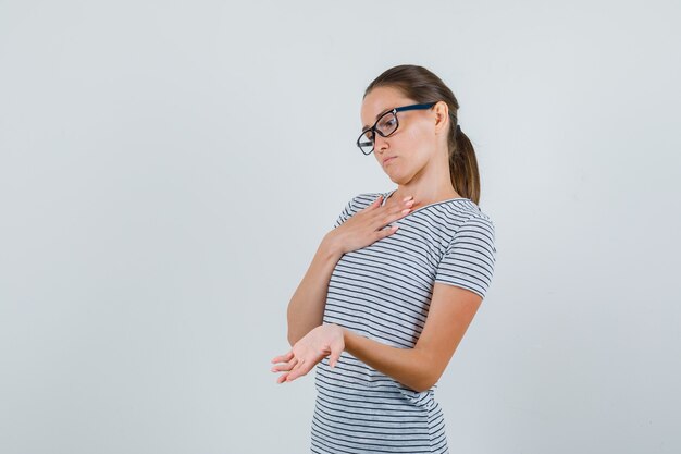 Mujer joven mostrando la palma en camiseta a rayas, gafas y mirando pensativo. vista frontal.