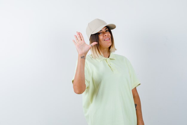 Mujer joven mostrando la palma de la mano para saludar en camiseta, gorra y mirando alegre. vista frontal.