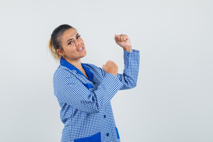 Foto gratuito mujer joven mostrando pose de ganador en camisa de pijama de cuadros azules y mirando bonita, vista frontal.