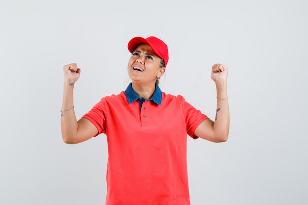 Mujer joven mostrando pose de ganador en camisa roja y gorra y mirando feliz. vista frontal.