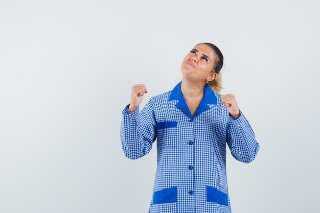 Mujer joven mostrando pose de ganador y mirando arriba en camisa de pijama de cuadros azules y mirando alegre, vista frontal.