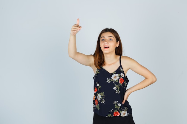 Foto gratuita mujer joven mostrando el pulgar hacia arriba en blusa y mirando sorprendido, vista frontal.