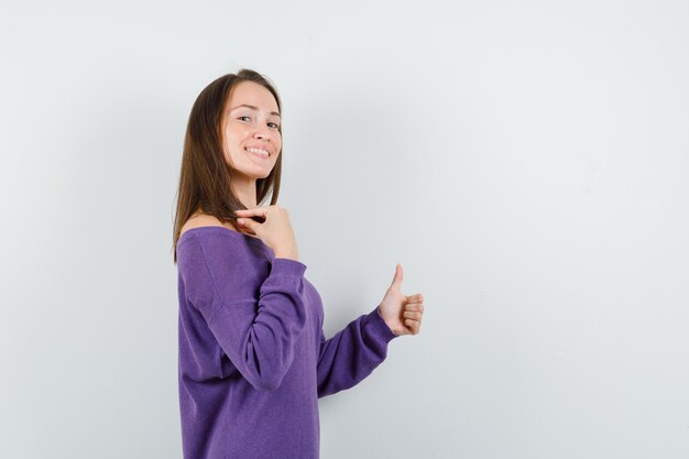 Mujer joven mostrando el pulgar hacia arriba en camisa violeta y luciendo optimista.
