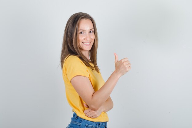 Mujer joven mostrando el pulgar hacia arriba en camiseta amarilla, pantalones cortos y mirando alegre