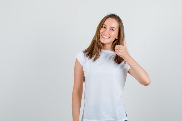 Mujer joven mostrando el pulgar hacia arriba en camiseta blanca y mirando feliz. vista frontal.