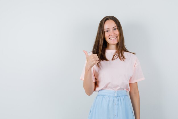 Mujer joven mostrando el pulgar hacia arriba en camiseta, falda y mirando alegre, vista frontal.