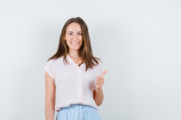 Mujer joven mostrando el pulgar hacia arriba en camiseta, falda y mirando alegre, vista frontal.