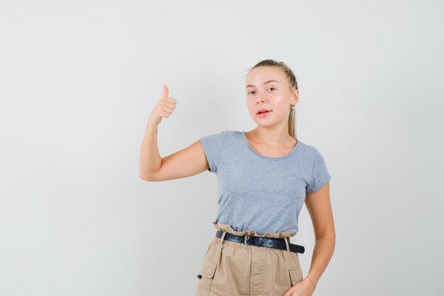 Mujer joven mostrando el pulgar hacia arriba en camiseta, pantalones y mirando alegre, vista frontal.